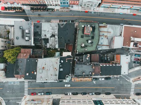 An aerial view of various rooftops, a street with cars, and pedestrians walking on pavements